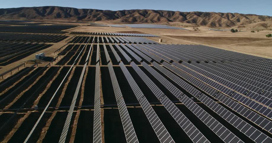 The sun shines brightly on solar panels on a solar farm surrounded by mountains