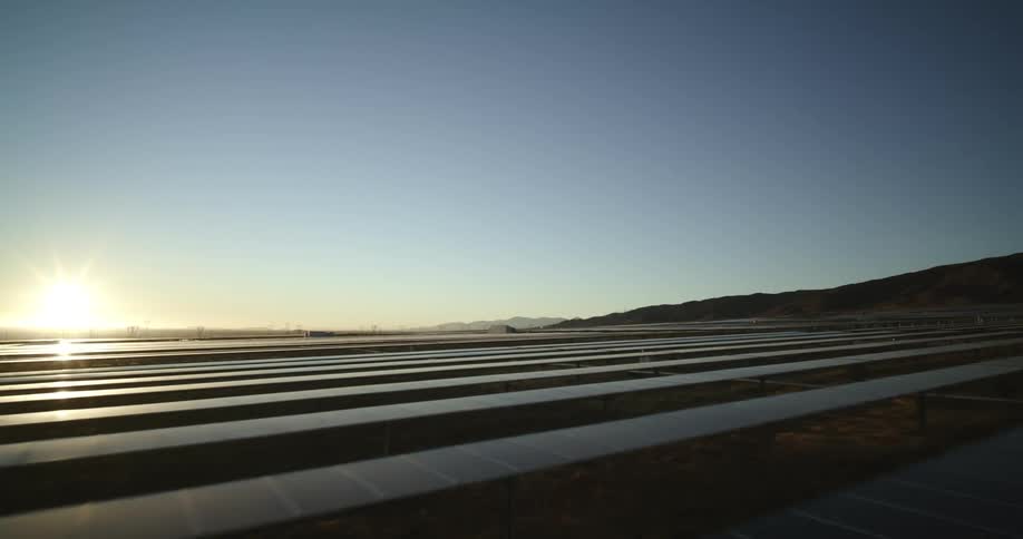 The sun shines brightly on solar panels on a solar farm surrounded by mountains