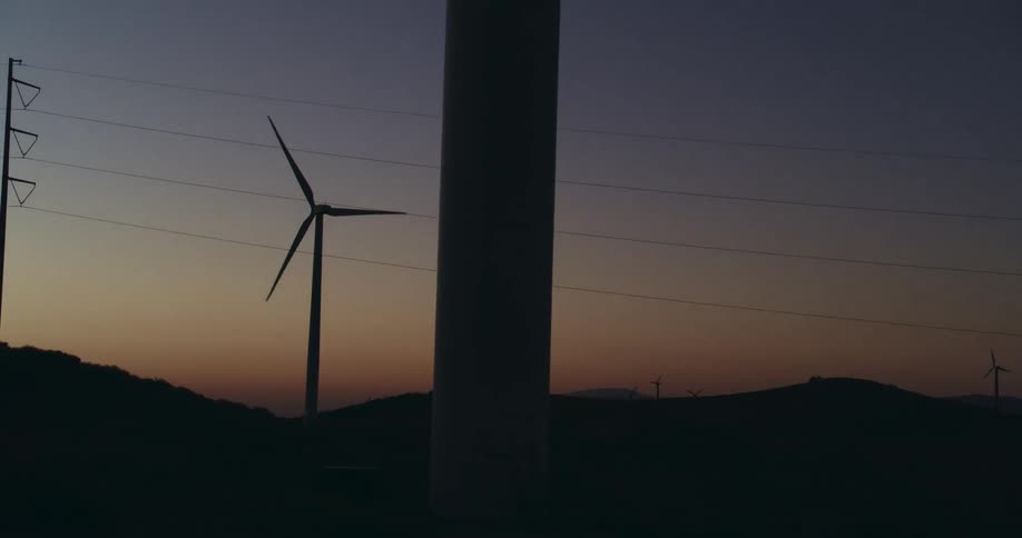 Wind turbines are spinning on a wind farm near utility wires in a darkening red sky