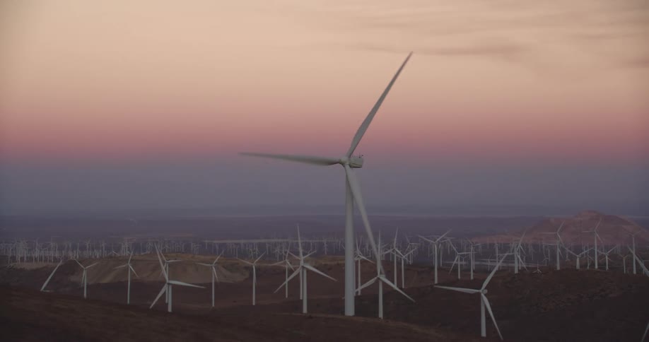 Wind turbines are spinning on a wind farm along hills with mountains in the distance under a pink sky