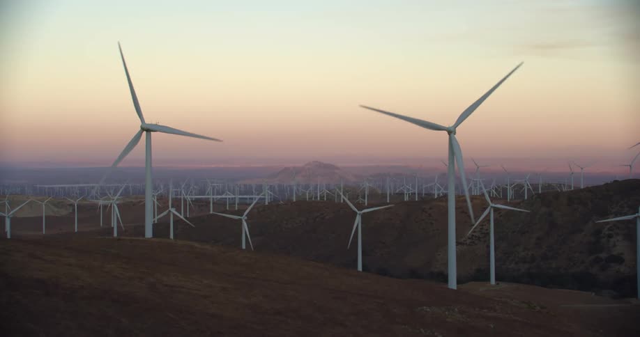 Wind turbines are spinning on a wind farm along hills with mountains in the distance under a pink sky