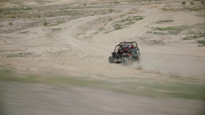 Four people in a dune buggy travel on a dirt road then go off the road into the desert as mountains sit in the distance under a blue sky