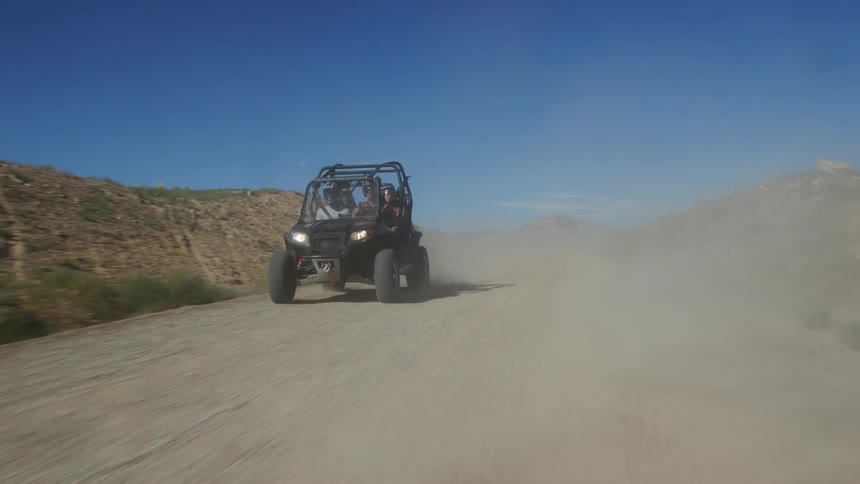 Driver and three passengers travel in a dune buggy on a dirt road as one passenger sticks his arm out and waves