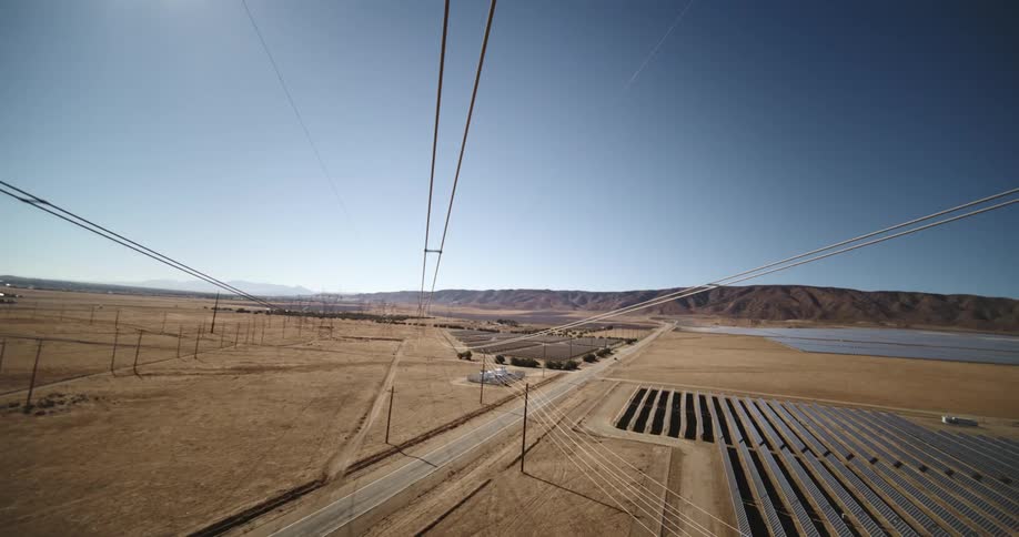 Utility towers and power lines are in open land with a solar farm and mountains in the distance