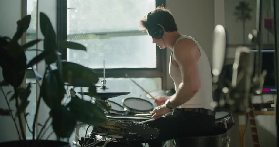 Man wearing headphones playing drums inside his house next to a glass window