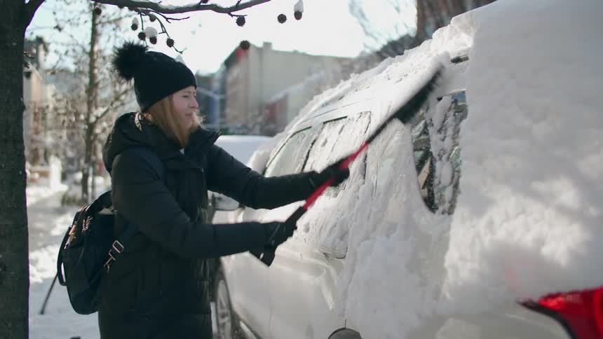 Woman wearing a jacket and winter beanie using a ice scraper to remove the snow covering a car in a public street
