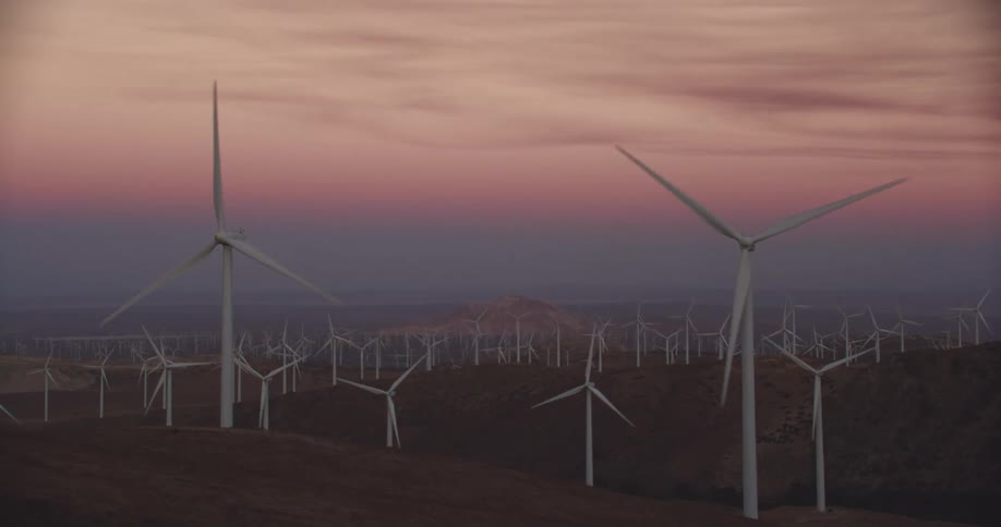 Wind turbines are spinning on a wind farm along hilltops with mountains in the distance under a red sky