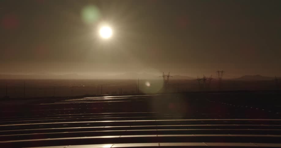 The sun shines brightly in a darkening sky on solar panels on a solar farm with mountains on the horizon