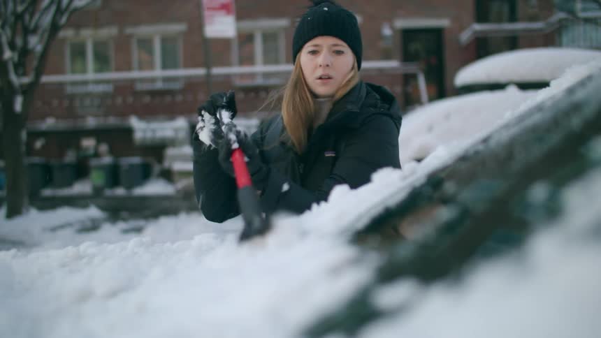 Woman in black winter jacket scraping snow off her car windshield