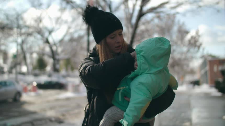 Worried mother wearing winter clothing carrying her baby wearing a green baby jacket down a street during winter