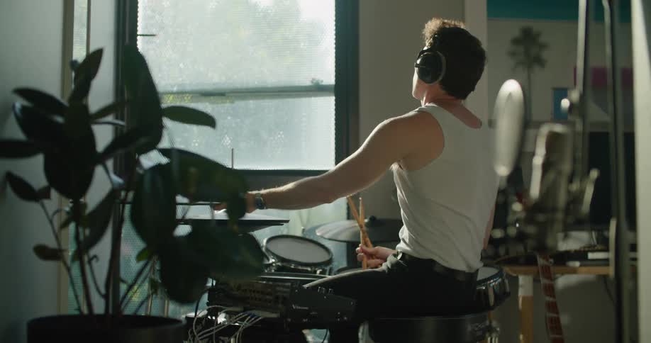 Young man wearing headphones playing drums in the living room at home in the morning