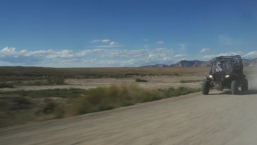 Dune buggy is being driven recklessly on a dirt road next to a field with mountains in the distance under a blue sky