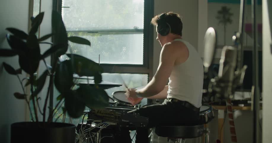 Man wearing headphones playing drums inside the house next to a big glass window