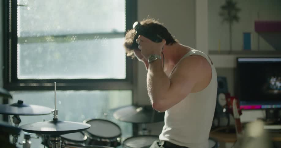 Man walks towards his drums sits and puts on his headphones and starts to play the musical instrument next to a big window