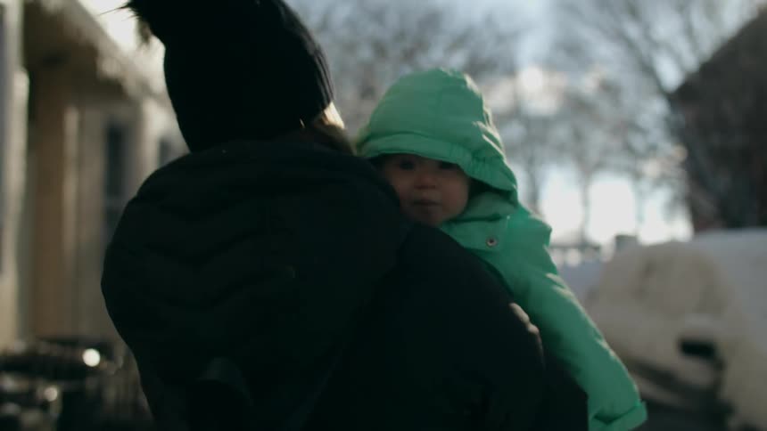 Mother wearing winter clothings walking down the snow covered street carrying her baby on her arms