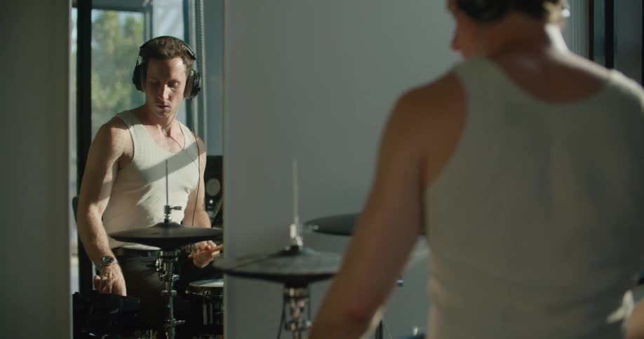A man at home sits down at his drums and puts headphones on and starts drumming