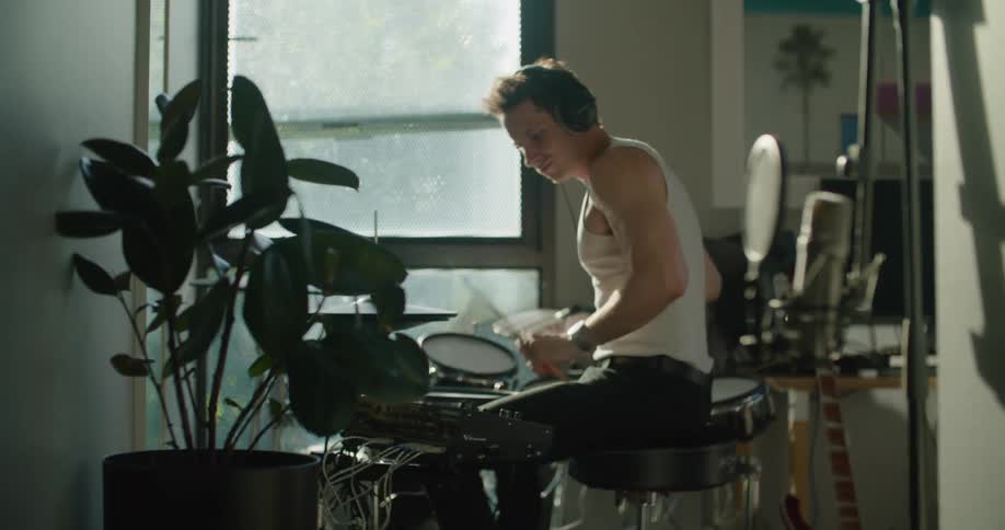 Male drummer with headphones white under shirt and black pants sits down and starts playing drums