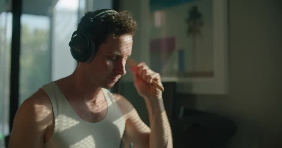 Man with white shirt and black headphones plays drums as the sun shines through windows