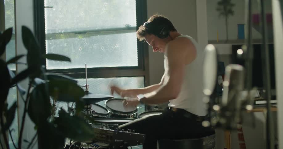The camera pushes towards a man playing drums indoors next to a big window