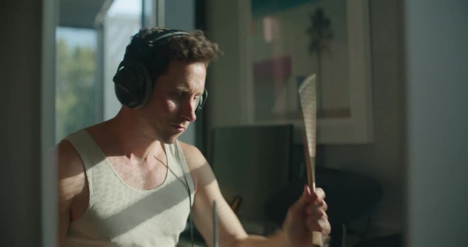 Man with white shirt and black headphones plays drums as the sun shines through windows