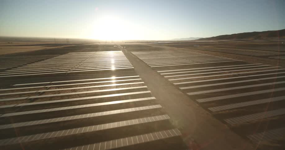 The sun shines brightly on solar panels on a solar farm with mountains in the distance