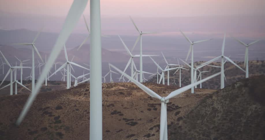 Wind turbines are spinning on a wind farm along hilltops with mountains in the distance under a pink sky