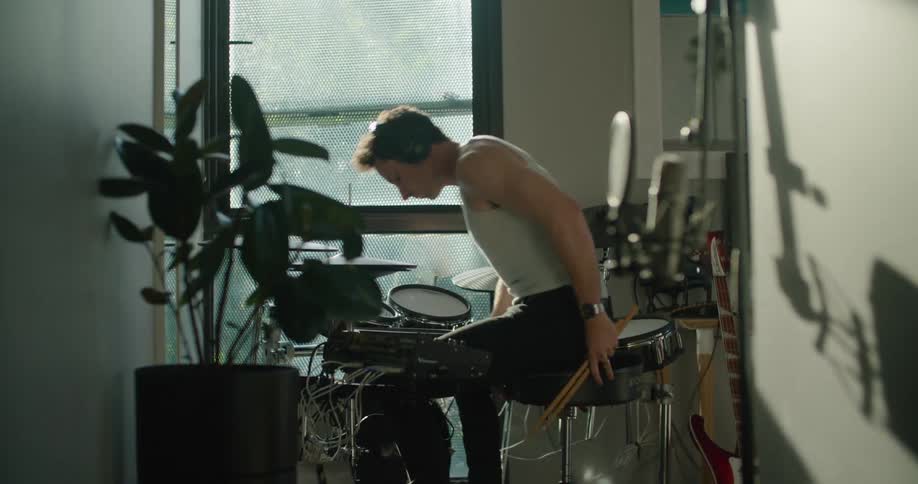 Man with white shirt and black headphones plays drums as the sun shines through windows