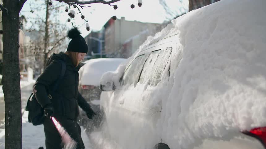 Woman wearing black jacket scraping fresh snow off her vehicle in a small town