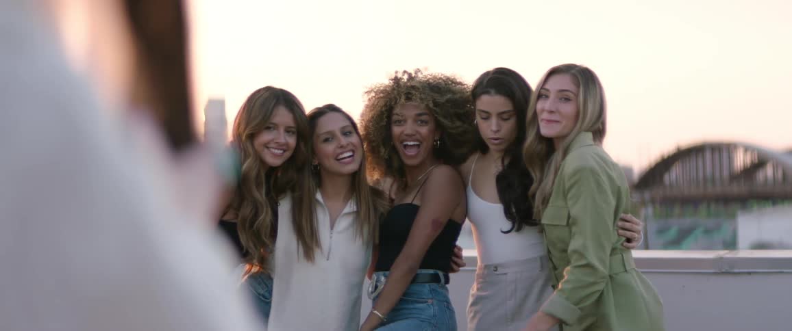 Five female friends smiling laughing and posing for a picture taken by a smartphone in front of a view of a citys skyline