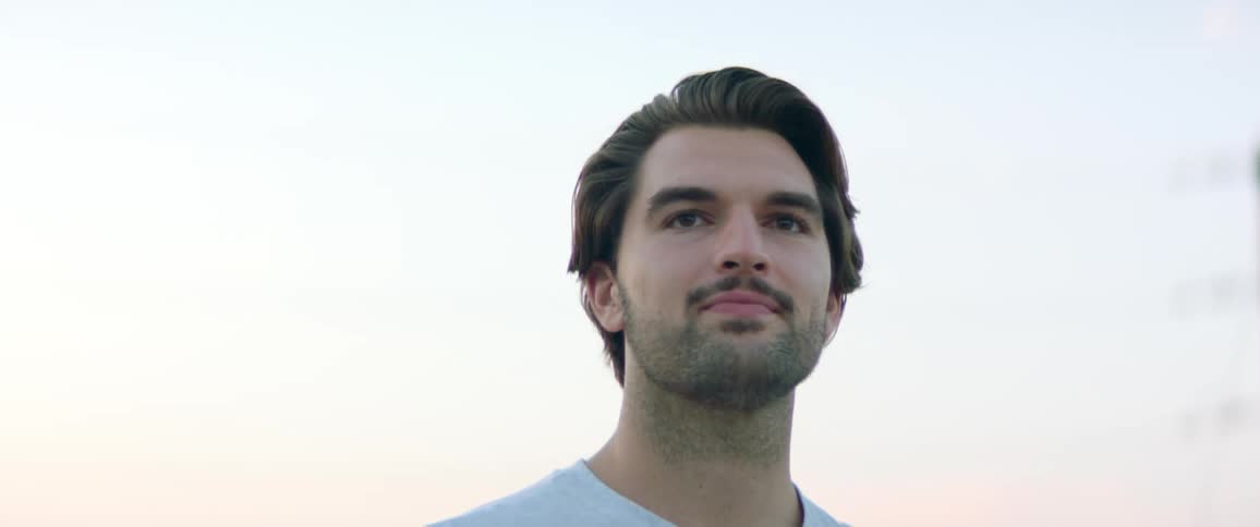 Man with a mustache and beard stubble stands outdoors near a post and looks thoughtful