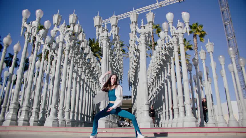 Female dancer dances near artistic display of rows of streetlamps as pedestrians move around