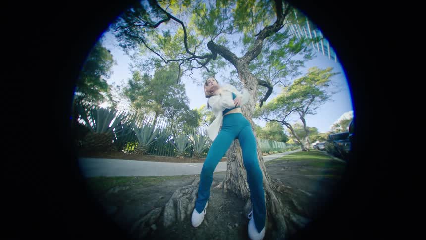 Young woman energetically dancing in front of a tree in a park at morning through a fisheye lens