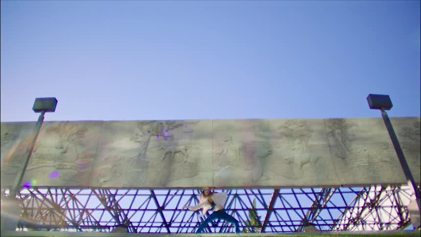 Woman wearing a blue top and white shirt dances in front of cement murals under a blue sky