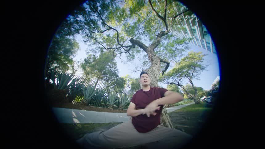 An asian male looks directly at the camera as he performs a dance outdoors under some trees