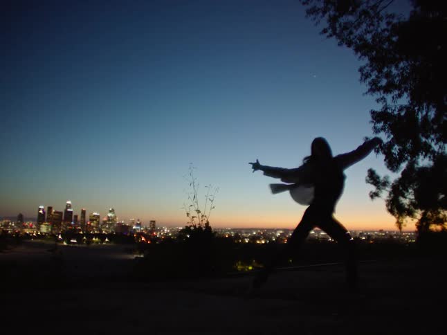 Dancing woman wearing shirt and trousers is silhouetted against sky and city skyline at dusk