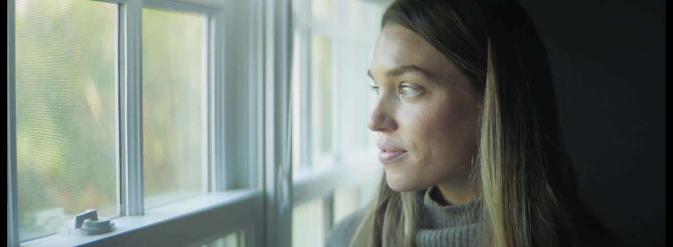 Pregnant woman with blonde hair and gray sweater looks out the windows in the kitchen