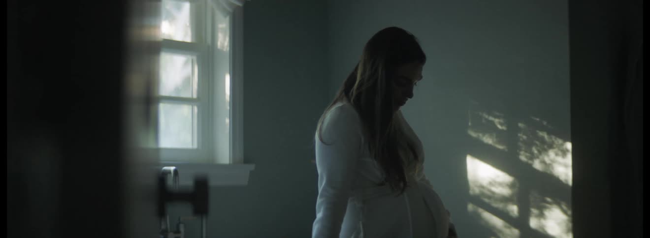 Pregnant woman with dark hair and white dress caresses her belly in the kitchen