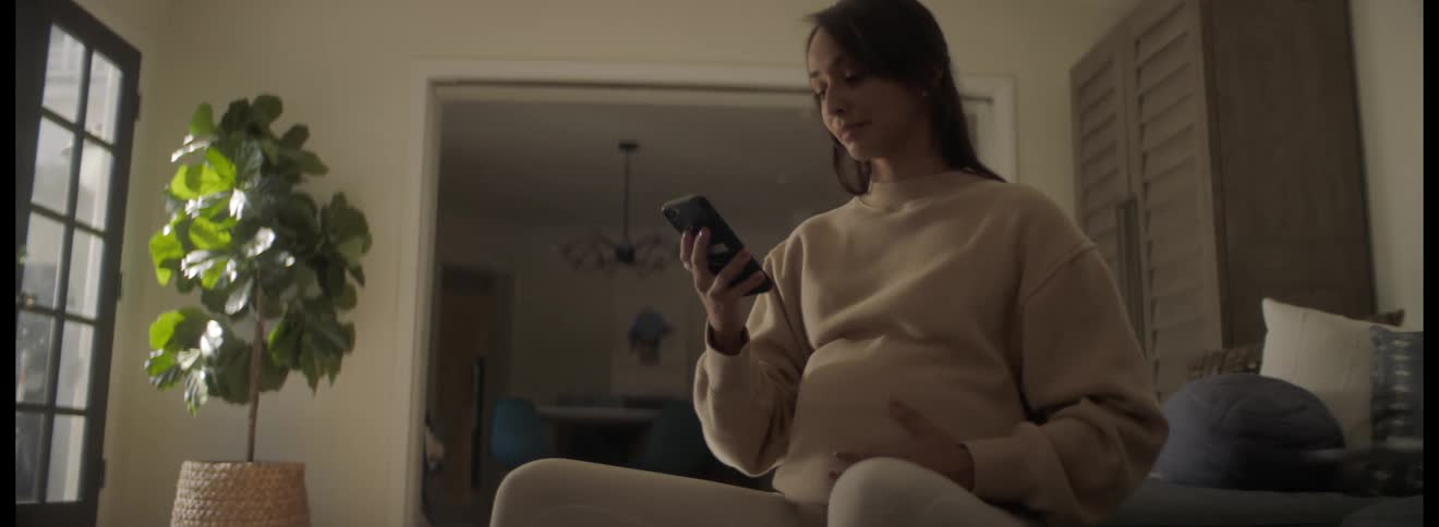 A pregnant woman sits on a stability ball at home and rubs her belly as she looks at her cell phone and smiles