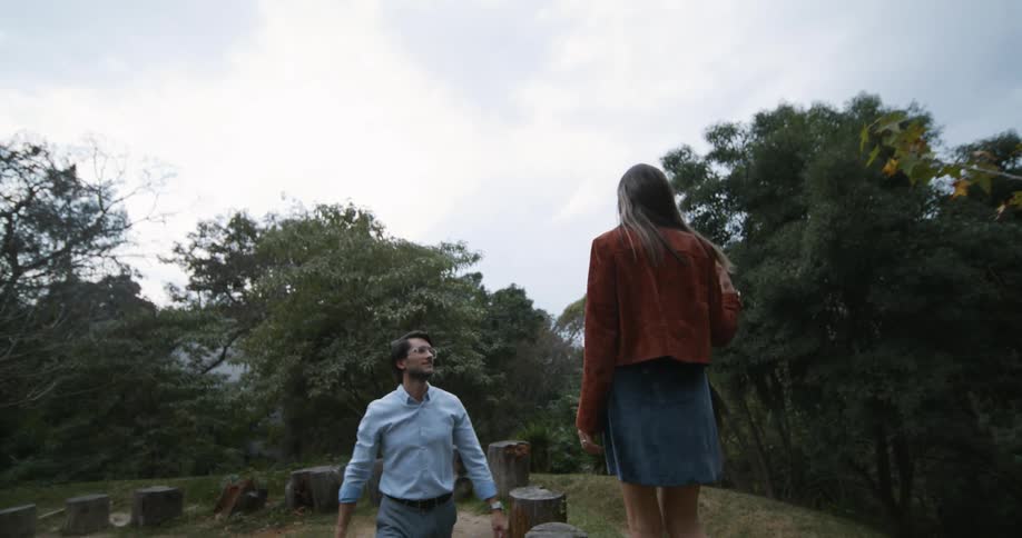 A man follows along as his girlfriend walks on tree stumps in a pattern in a park