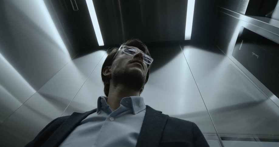 A man wearing glasses and a suit coat stands inside of an elevator and waits patiently