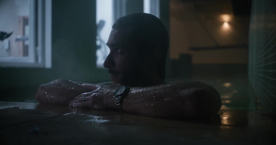 A man wading in warm water indoors approaches the edge of the shallow pool and props his arms on it