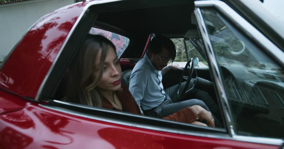 Young sad depressed woman sitting on a red sports car while a man sits on the driver seat