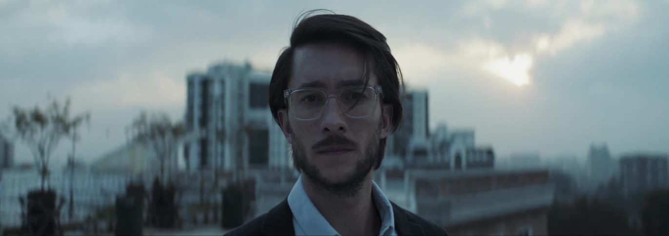 Man with glasses beard shirt and blazer jacket faces the camera on rooftop with buildings in the background at dusk