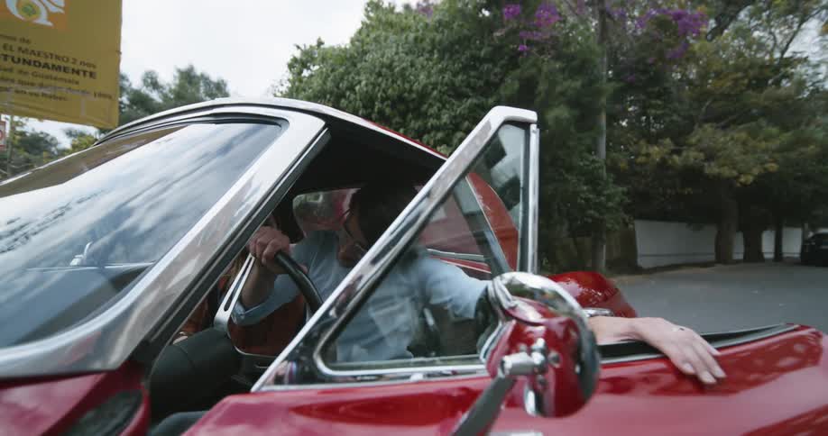 Man enters a red vintage car and sits at the drivers seat while a woman awaits at the passenger seat