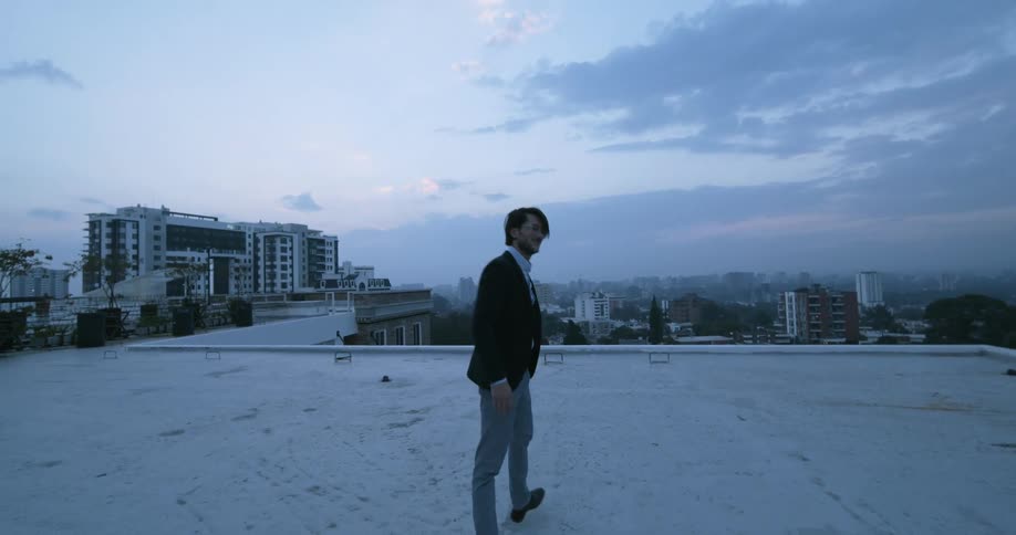 Young man wearing glasses walks away from the camera on the roof of a building at sunset