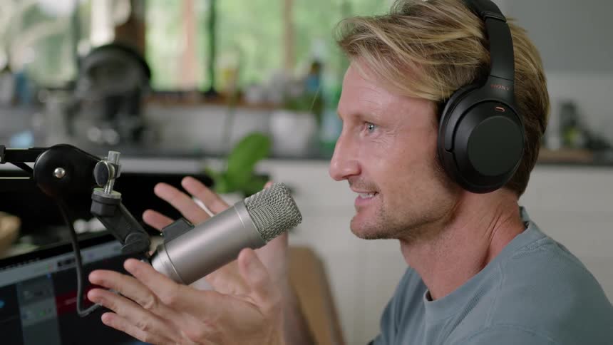A man wearing headphones is sitting down and moving his hands as he speaks into a microphone