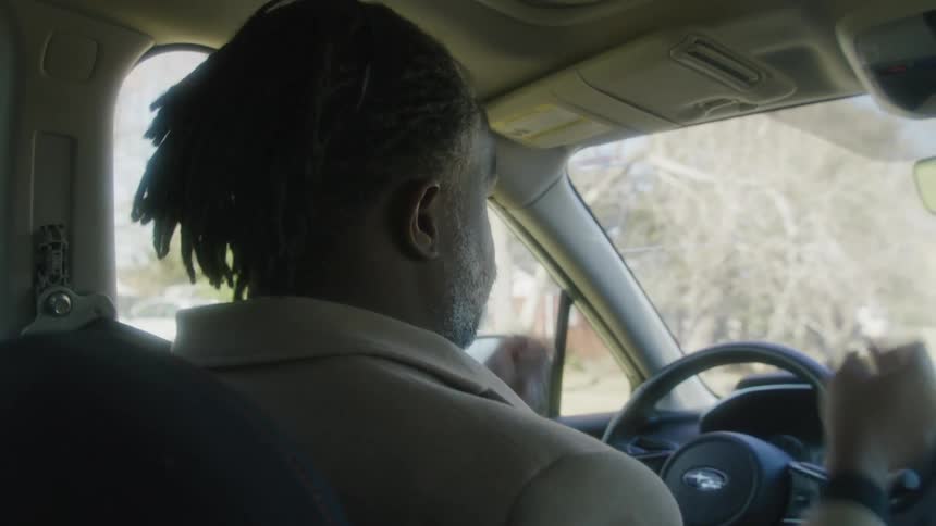 Man with dreadlocks beard and brown overcoat sits in car and shakes his head in disappointment