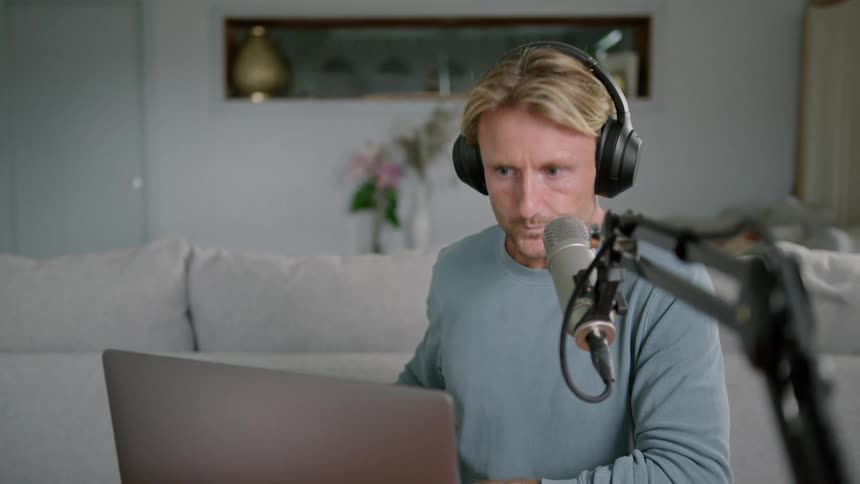 Man wearing headphones drinks from a coffee mug then smiles and talks into a microphone as he looks at his laptop
