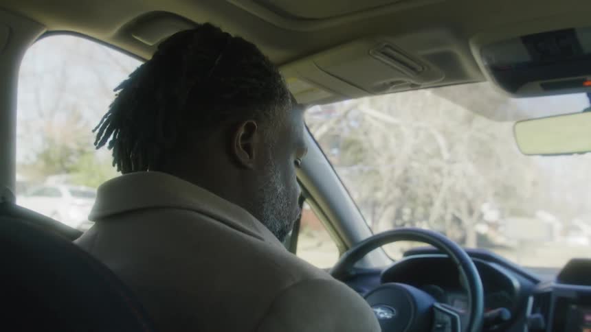 Man with dreadlocks beard and brown overcoat drives and stops the car shaking his head