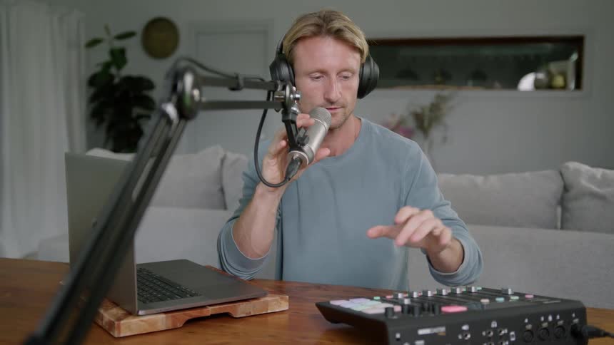 A man sitting in front of a laptop at home puts on headphones and talks in to a microphone